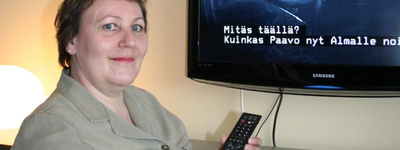 Nainen katsoaa tekstitettyä televisio-ohjelmaa kaukosäädin kädessään.