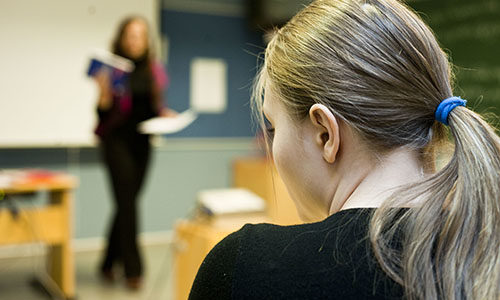 Nainen istuu luennolla, taustalla opettaja.