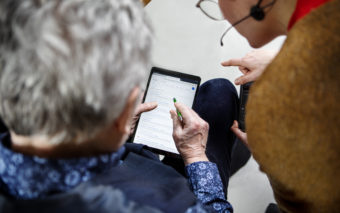 Kaksi henkilöä ovat tabletin äärellä. Vasemmalla herrasmies katsoo tablettia digikynä kädessään. Oikealla seisova henkilö neuvoo.