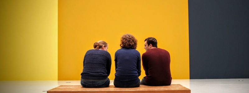 Kolme ihmistä istuu puisella penkillä näyttelytilassa. Taustalla keltainen ja harmaa seinä.