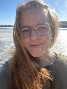 Jenni Nuutisesta kuva, ruskeat hiukset, silmälasit ja vihreä paita. Taustalla näkyy jäätynyttä järveä ja aurinkoa.