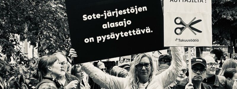 Ihmisiä järjestöjen mielenosoituksessa Esplanadin puistossa Helsingissä. Kuuloliiton toiminnanjohtaja Sanna Kaijanen pitelee kylttiä, jossa lukee ''Sote-järjestöjen alasajo on pysäytettävä.''