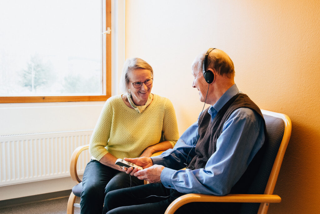 nainen katsoo hymyillen vastapäätä istuvaan mieheen, jolla kommunikaattori käytössä